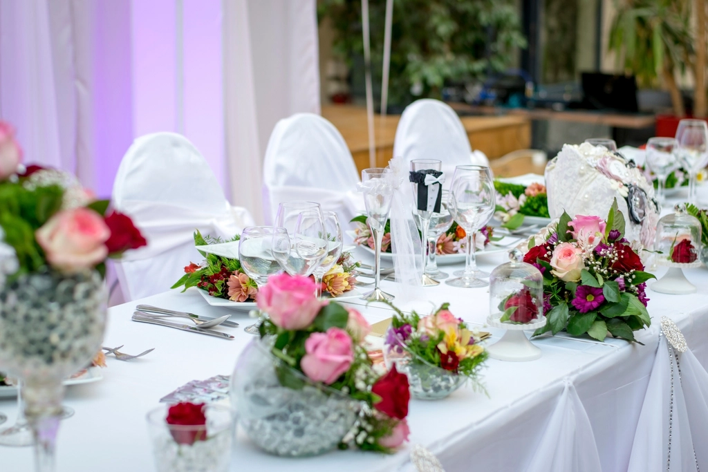 Wedding reception table dressed with pink and red roses, crystal glasses and white chair covers